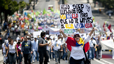 An opposition demonstrator with a Venezuelan national flag draped around her shoulders, holds a sign with a message that reads in Spanish; "Son, I keep fighting for Venezuela, so that you don't leave it, or sink with it," during a May Day march in Caracas, Venezuela, Friday, May 1, 2015. Pro-government and opposition workers marched today in the midst of the political and economic crisis prevailing in the country, with separate marches that took place to mark International Workers Day also known as May Day. (AP Photo/Ariana Cubillos)